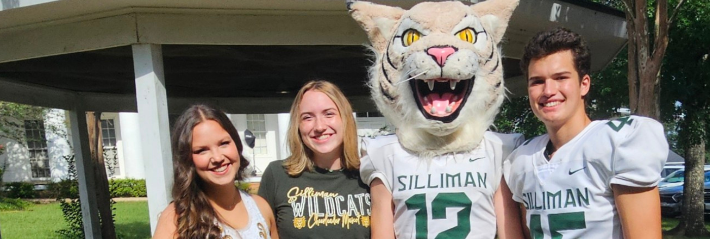 Image of four students posing in front of the school. One student is dressed as Willie the Wildcat.